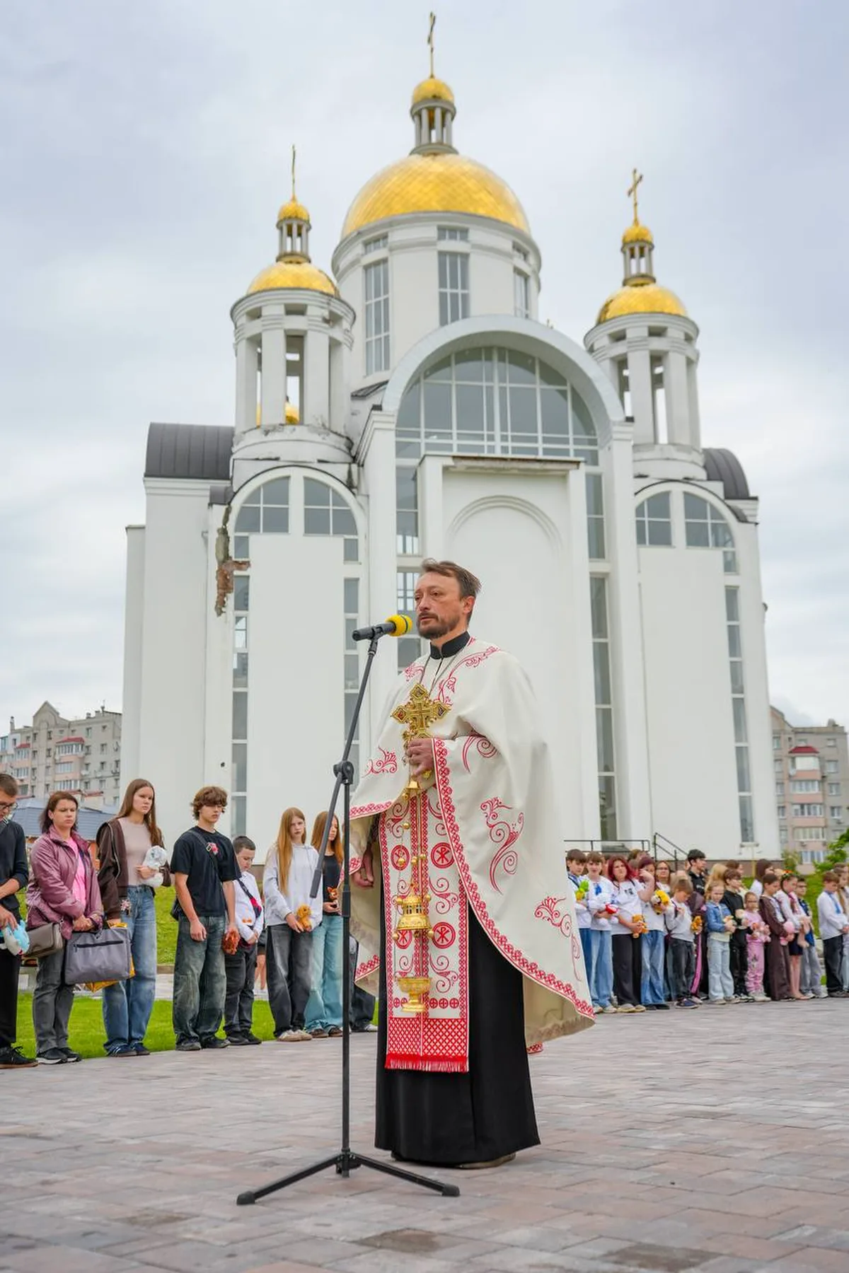 Священик у церковному облаченні перед білою церквою з золотими куполами