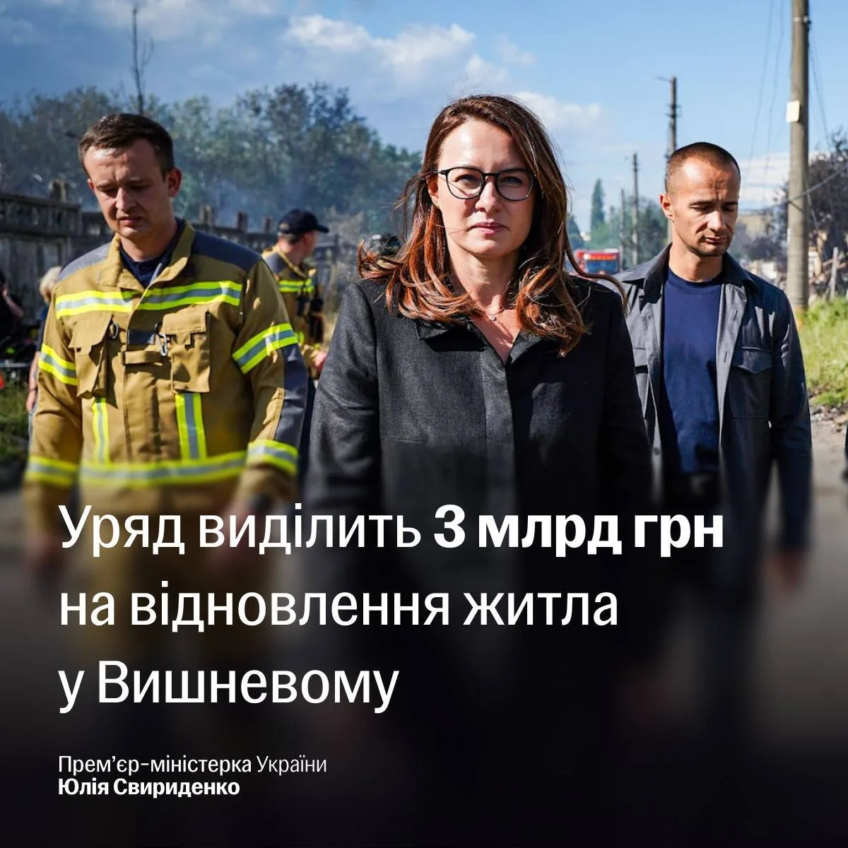 Жінка в окулярах та чорному піджаку з рятувальниками на фоні