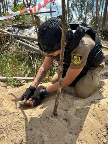 Піротехнік у захисному спорядженні обстежує знайдений боєприпас у грунті