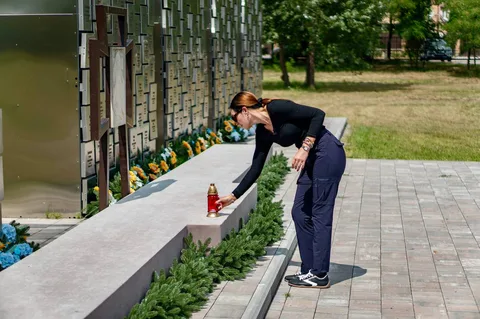 Жінка в темному костюмі схиляється до меморіальної стіни з квітами