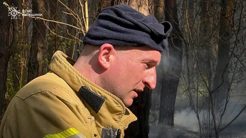 Чоловік у жовтій куртці та чорній шапці біля палаючих дерев